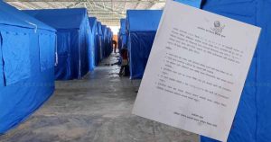 Rows of temporary shelters inside a holding centre with an official notice displayed in the foreground